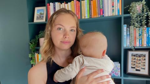 A woman with blonde hair and blue eyes looks at the camera while holding her baby, who faces away from the camera. In the background are blue shelves and books. 