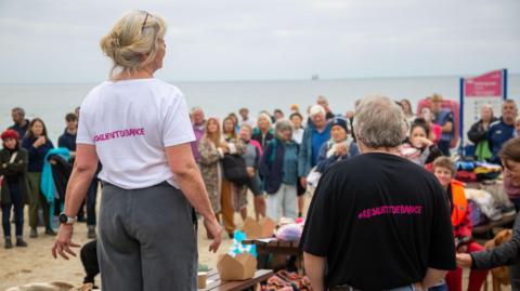 Ruth Hitchcock and Dave Hitchcock with their backs to the camera and facing a crowd of people. They are wearing t-shirts which say #resilientdefiance on the back in pink letters. The sky is grey. The sea is a light grey. 