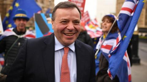The image shows Arron Banks in Westminster walking away from pro-EU campaigners. He is smiling and wearing a dark suit, light blue shirt and red tie.