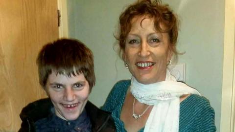 A boy with dark hair is smiling. He's with his mum who is wearing a blue top and white scarf. She has a necklace and is also smiling. There's a door behind them and a blue wall. The boy has a dark coat and top on.