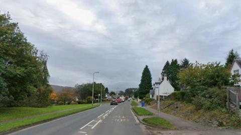 The Road to the Isles at its entrance to Corpach, in Lochaber. Skies are grey, there are trees on either side of the road and some houses on the right. There is a bus stop in the frame.