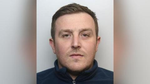 A mugshot of a man who has his brown hair combed over to one side. He has facial hair and is wearing a blue sweater.