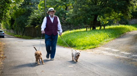 Robin Shuckburgh on a country road walking his dogs, Gizmo and Widget. They are walking by a green space. It is a sunny day.