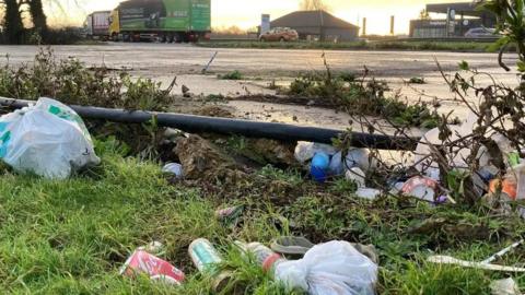 Rubbish including drink containers strewn along a stretch of the A1. Vehicles can be seen in the background, along with a number of buildings.