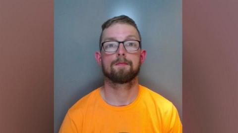 A mugshot of a man with short, brown hair and glasses wearing a bright yellow t-shirt.