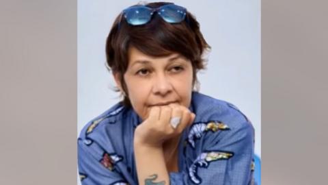 Karine Buisset, who has her hand on her mouth. She is wearing a blue top and has medium-length brown hair with sunglasses on her head.