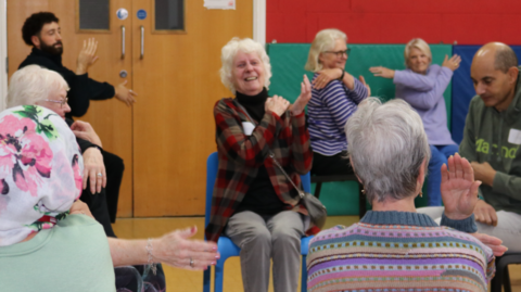 Eight people are in a room and some of them have their hands near their face. Doors are on the left of the photo.