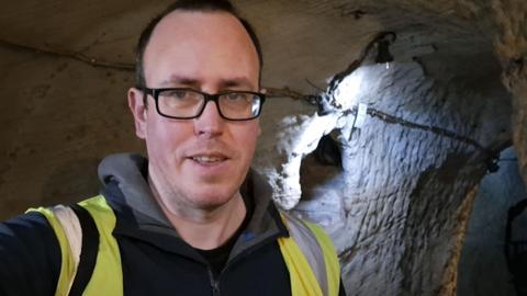A view of a man with glasses and a hi vis vest stood in a grey cave.