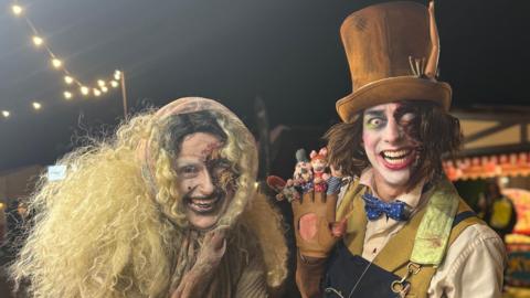Two horror characters smile at the camera. Phoebe is on the left wearing a blonde curly long wig and dressed as a witch with blackened teeth and a scarred face. Next to her is a Mad Hatter character with one missing eye and an albino eye. He wears a mustard top hat, blue bow tie and glove with mini puppets on and has a maniacal smile. Behind are fairground rides.