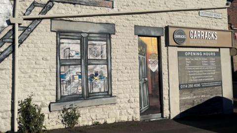 The exterior of bricked building. There is a large mural of a window  painted in a realistic style. To the right of the window mural is a painted door featuring a portrait of an older person wearing glasses and a hat. Above the door is a black and white sign that reads 'Carracks Wallpaper & Paint'.