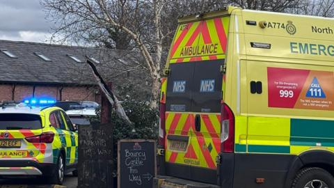 An ambulance and ambulance response vehicle are parked at the farm. A sign says 'bouncy castle this way' while another says 'parking this way'.