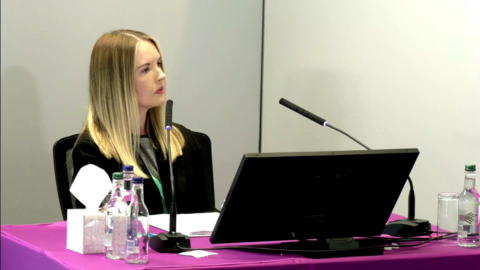Service manager Chloe Cawston sits in front of a table at the Lampard Inquiry. She is wearing a black jacket and a grey top underneath it. There are two microphones pointing torwards her and a laptop in front of her, to each side there are bottles of still and sparkling water and a box of tissues, the walls are white, the tablecloth is purple and she has long blonde hair.