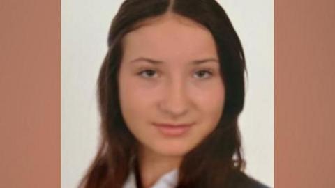 A teenage girl with long brown hair looks at the camera. She is wearing a white shirt, a navy and white striped tie and a navy blazer.