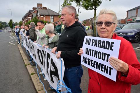 Woman wearing red jacket holds a sign which reads no parade without agreement