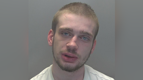 Custody image of Moorcroft against a grey background. His head is leaning slightly to the right and he has light brown facial hair and short hair. His eyes are blue and he is wearing a white top. 