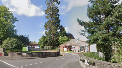 A street view shot showing the entrance to HMP Leyhill, it has a small entrance building and a small road leading to a car park with trees on either side