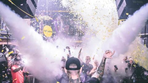 The picture shows a packed nightclub scene during a high‑energy moment in the middle of a DJ set. The photo is taken from behind the DJ booth, with the DJ in the foreground facing the crowd. Smoke cannons fire bursts of white fog across the room, and yellow confetti fills the air. People in the crowd are holding up their phones, filming the atmosphere and the performance. Lights, speakers and balconies full of people frame the scene, and the venue logo “THE DEPØ” appears in the bottom right corner.
