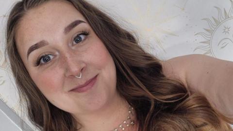 A smiling woman with shoulder length light brown hair. She is taking a selfie photo and has bare arms, a nose ring and a silver necklace
