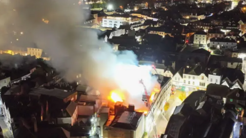 A picture of the fire at the former Debenhams store in Canterbury, captured from a bird's eye view. Two cranes erected over the roof are trying to put the fire out, and smoke is billowing across the nearby area.