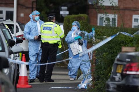 Police and forensics officers stand outside a house in Uxbridge. The area is cordoned off with police tape and traffic cones as officers guard the scene.