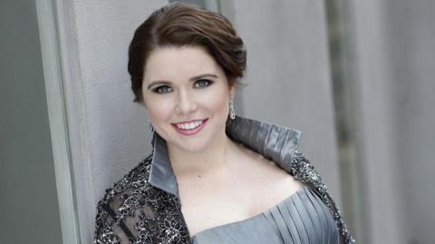 Tara Erraught stands in front of a grey wall and is smiling. She is wearing a silver formal dress and silver earrings, her hair is tied up