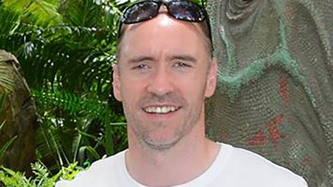 Paul Doyle has short dark greying hair with sunglasses on his head and is wearing a white T-shirt.