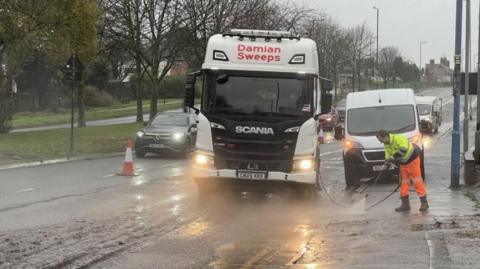 A lorry in a flooded street with a car passing by on the other side of the road, past an orange traffic cone, with a man with a hose attempting to clean the roadside