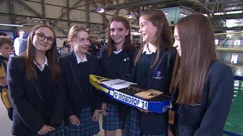 Five girls standing smiling at the camera, wearing school uniform and holding a blue and yellow mini speed boat, which has 'Snowdrop '24' written on it.