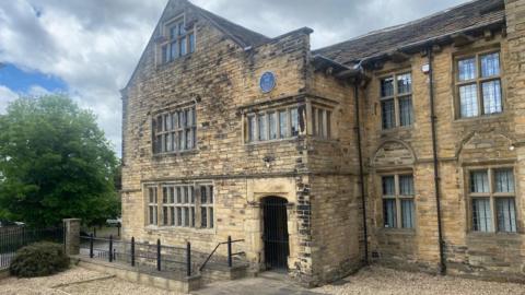 A 17th Century sandstone building with stone mullion windows and a blue plaque above a first floor window over the doorway. To the left is a mature tree and there is a cloudy sky with patches of blue.