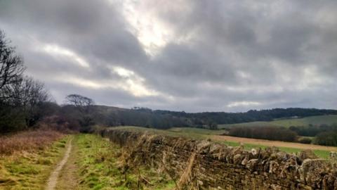 Sky filled with thick grey clouds and a few brighter breaks, over fields and a wall edging a field