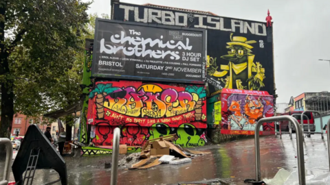 Photo of Turbo Island area. A building with graffiti and a black and white billboard. Park cars surrounding the area with bike racks. In the middle of the small area there is burnt debris and cardboard in a pile. 