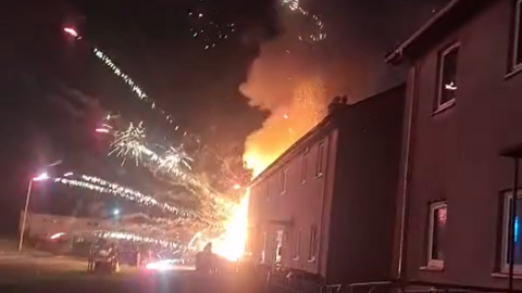 Fireworks explode from a fire obscured by a row of houses on Braes Avenue. A cloud of orange smoke rises above the buildings. The fireworks are shooting across the night sky into an open space opposite the homes. 