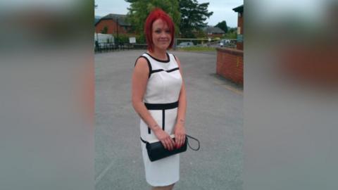 Tiffany Render has dyed-red hair and is smiling. She is standing near a building, dressed in a smart long white and black dress holding a black handbag.