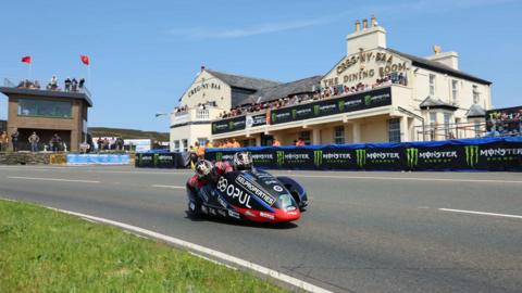 A black and red sidecar outfit travelling past the Creg ny Baa, which has crash barriers in front of it.