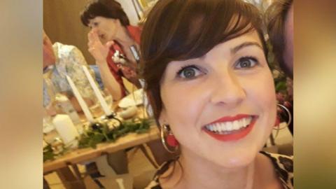 A woman with dark hair and wearing hoop earrings smiling at the camera. There's a table behind her with some candles on, and a woman sitting at the table mid-conversation.