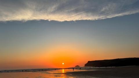 A picture of the sunrise over the beach. There is a large beach and sea with rocks in the distance. There is a orange skyline.