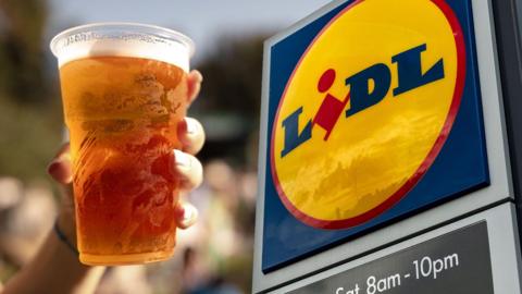 A graphic showing a hand holding a plastic pint glass of beer in front of a yellow, blue and red Lidl sign. 