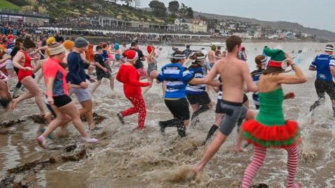 People in fancy dress running into the sea with water splashing up and crowds lining a beach.