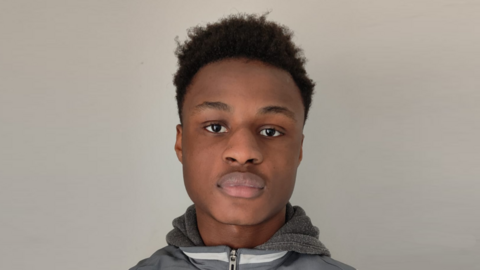 A man with dark hair, wearing a hoodie, pictured close-up to his face with a plain white wall behind him