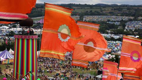 Six large orange festival flags fly in the wind. In the background is a tall ribbon tower, which is in one of the field's at Glastonbury festival. Crowds of people can be seen underneath it, with thousands of tents in campsites as far as the eye can see.