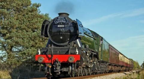 The Flying Scotsman - an impressive black, green and red steam locomotive. It is alongside a green hedgerow and is pulling burgandy and cream coloured carriages. 