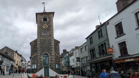 A general view of Moot Hall in Keswick. It is in the middle of a pedestrian area and has a turret with two sets of steep steps in front of it, each leading up to the central doorway. The brick building also has a black clock with golden numbers and features. There are black and orange table and chairs from a cafe outside the building.