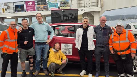 A group of seven men stand in front of a car on a vehicle ferry. They are side by side and are smiling at the camera. They are standing in front of their red car. The water and boats are visible behind them. 