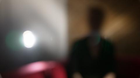 Emma sits indoors, deliberately photographed heavily out of focus so that her face and identifying features cannot be seen. A bright light is visible to the left and a red sofa is behind her.