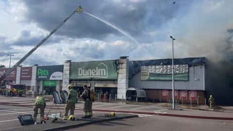 Retail park showing various units including Dunelm and Pets at Homes. The charred interior of the Dunelm shop is visible. One fire crew is directing a jet of water from above down into the smoke-filled Dunelm unit. Some firefighters are looking at a plan of the site in the foreground