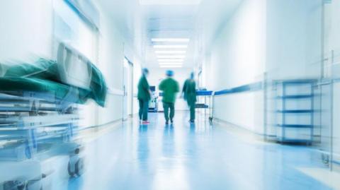 Stock image shows a blurred hospital ward. In the distance you can see the figures of three people wearing scrubs and hats.