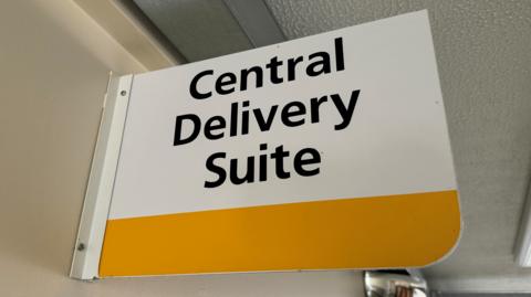 A white sign with black writing saying Central Delivery Suite. There is a blank yellow strip underneath the writing for design purposes. 