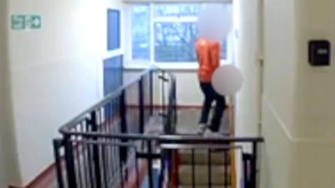 A person standing at the top of a flight of stairs. He is wearing a red polo shirt, dark trousers and white trainers. His face and groin are are blurred out.