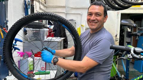 A dark-haired man in a blue T-shirt, smiling and holding a bike wheel in a workshop.