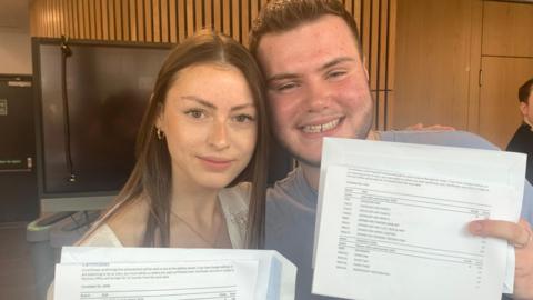 A female student with long brown haor and a male with short brown hair hold up their exam results. 